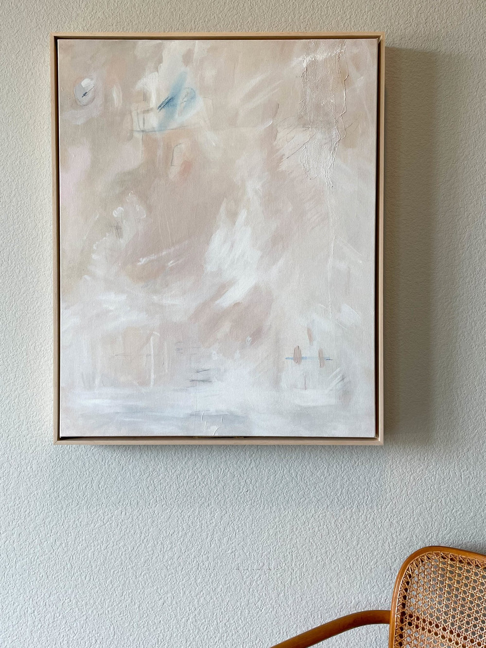 Light abstract acrylic painting with pops of soft blue by Ashley Keane Familiar Soul with maple frame hung on wall above wicker chair - Artly International