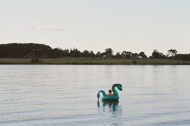 Photograph of woman floating on lake near a lake house in Maine - Artly International